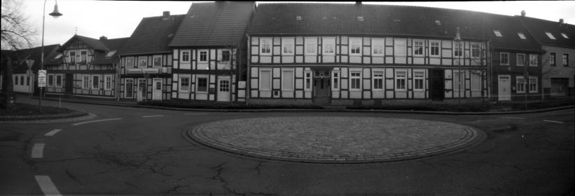 Schwarzweiß Panoramaaufnahme.
Fachwerkhäuserzeile, davor ein Minikreisverkehr.