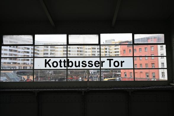 View from inside the elevated Kottbusser Tor U-Bahn station in Berlin, looking out through a window with the station name "Kottbusser Tor" printed in bold black letters. Behind the glass, a dense urban scene is visible with modernist residential buildings, older red and white facades, storefronts, and street-level activity below.