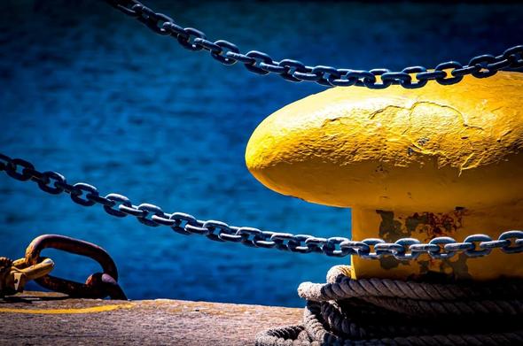The image shows a large yellow bollard on a pier or dock, with thick ropes and chains secured around it. In the background, there is a body of blue water, suggesting a maritime setting.