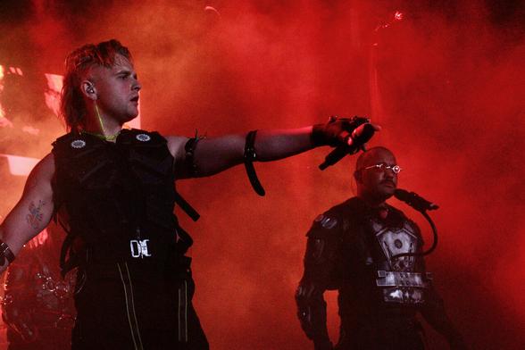 Two members of a band performing; red lighting, stage smoke.