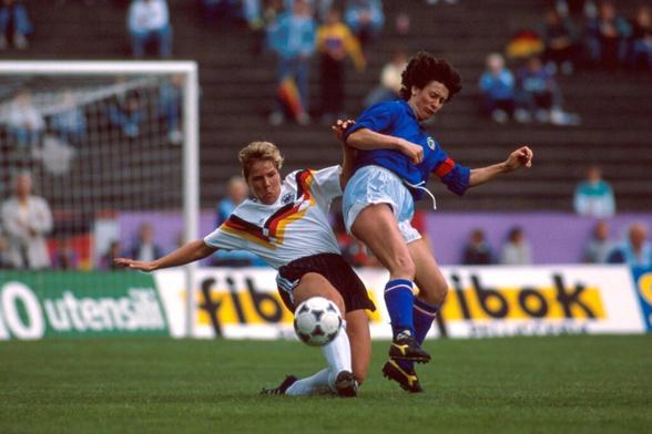 Deutschlands Verteidigerin Frauke Kuhlmann im Zweikampf mit Italiens Stürmerin Elisabetta Vignotto. Kuhlmann links im Bild kniet mit einem Bein auf dem Rasen und macht mit dem anderen einen Spreizschritt, um den Ball wegzuspitzeln, sie schaut fokussiert auf den Ball und hat beide Arme von sich weggestreckt, um die Balance zu halten und Vignotto auf Distanz zu halten, die rechts neben ihr mit dem Rücken zu ihr nach einem Sprung in Richtung des Balles auf dem Boden zu landen scheint. Auch sie versucht noch ein Bein nach dem Ball auszustrecken, hat aber die ungünstigere Position, weil er schräg hinter ihr ist.