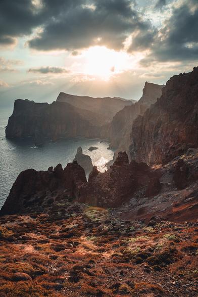 Dramatic coastal landscape with rugged, reddish-brown rock formations jutting into the sea. In the background, the sun shines through dense clouds, casting a warm, golden light over the scene. The calm sea gently reflects the sun's rays.
