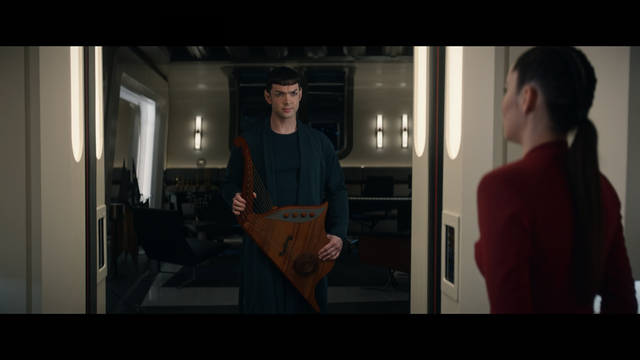 A Star Trek Strange New Worlds shot of Spock holding his Vulcan lute and standing in the doorway to his quarters as he talks with La'an on the Enterprise.  From E203