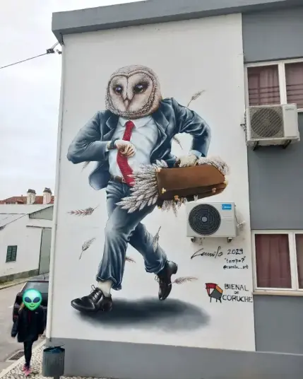 A mural painted on the side of a building depicts a surreal scene of a humanoid figure with the head of an owl. 

The figure is dressed in a business suit with a red tie and is carrying a briefcase from which feathers are spilling out. 

The mural includes the artist's signature, "Camilo 2023 'tempo?'" and the Instagram handle "@camilo_art". 

Below the signature, it says "BIENAL DE CORUCHE" with a small logo. 

An air conditioning unit is mounted on the wall next to the mural. In the foreground, a person with an alien emoji covering their face is walking past the mural.