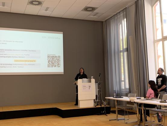 Lady at a lectern in front of slides. The three workshop organizers are visible, and I am SO jealous of their t-shirts 😍😍😍