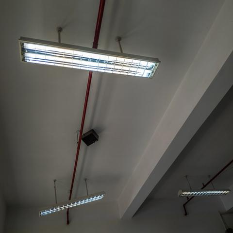 A picture of three tube light holders packed with tube lights hanging from the ceiling with red firefighting pipes running in between them. The lights are hanging on the ceiling with two anchor points. One of the tubelight holders in the background has lost one of its anchor points to the ceiling and is hanging on by a single one in a slanted manner.