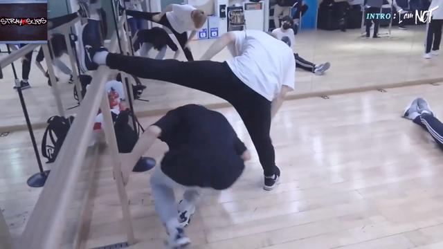 Felix is stretching in the dance practice room, with one leg up on a barre. I.N crouches down to run underneath Felix's leg.
