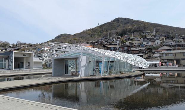 I have no idea how to even start describing this. Okay, in the background there is a traditional Japanese village on a hill. On the foreground, it's a white structure that vaguely resembles a seashell. It's made of metal but it's actually "lace" made of various scripts from various languages. oh and it's actually the roof of a building made of glass, that happens to be the welcome center of the island. Does it made any sense?