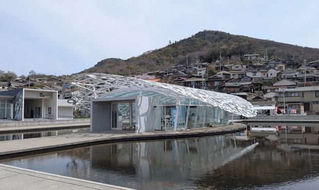 I have no idea how to even start describing this. Okay, in the background there is a traditional Japanese village on a hill. On the foreground, it's a white structure that vaguely resembles a seashell. It's made of metal but it's actually "lace" made of various scripts from various languages. oh and it's actually the roof of a building made of glass, that happens to be the welcome center of the island. Does it made any sense?