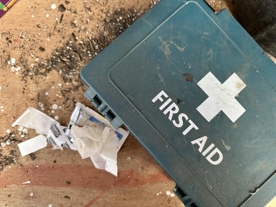 A first aid kit with a used sterile and the packaging from a plaster. They are sitting on a potting bench with some compost and perlite.