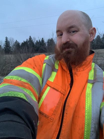 A large cis white male offers out a hug. He wears a reflective jacket and has a red beard