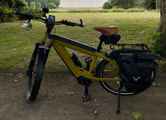 Das Bild zeigt ein modernes gelbes E-Bike, das auf einem grasbewachsenen Weg neben einem Baum geparkt ist. Das Fahrrad hat einen robusten Rahmen mit einem schwarzen Akku, der am Unterrohr montiert ist. Es verfügt über einen digitalen Tachometer am Lenker, breite Reifen und eine Federgabel, die auf Komfort und Stabilität hinweisen. Zusätzlich ist das Fahrrad mit einem braunen Ledersattel, einem Gepäckträger hinten und zwei großen schwarzen Packtaschen ausgestattet, die an den Seiten des Hinterrads befestigt sind. Die Umgebung ist grün und friedlich, mit Bäumen und Büschen im Hintergrund, was auf eine ländliche oder parkähnliche Umgebung hindeutet.