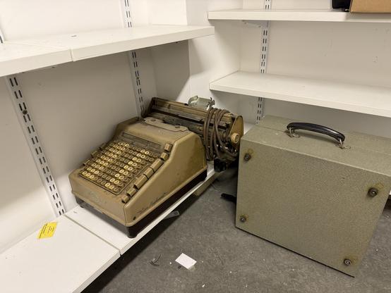An old adding machine is positioned on a shelf next to a gray carrying case. The typewriter is beige with yellow keys, showing signs of wear and age. The shelf has a few empty spaces, and the floor is visible beneath.