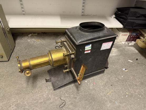 An antique machine with a metallic body and a cylindrical brass attachment in the front, likely a historical camera or projector. The device is located on a cluttered surface with various items in the background.