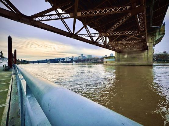 Burnside Bridge stretches across the Willamette River overhead, viewed from the floating pedestrian paths of the Willamette Greenway/Eastbank Esplanade on a cloudy afternoon.