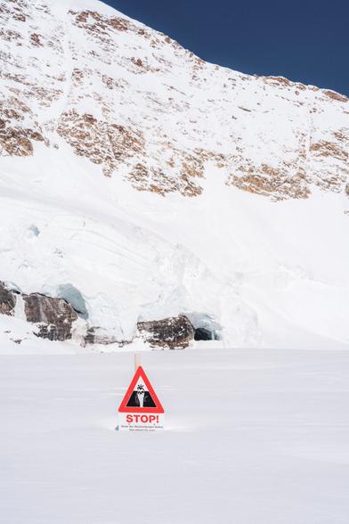 The image shows a snowy mountain landscape with a stop sign warning about potential avalanches. The sign features a triangular warning symbol with a pictogram of falling rocks or snow, and below it, the word "STOP!" in bold red letters. The background shows snow-covered rocks and cliffs.