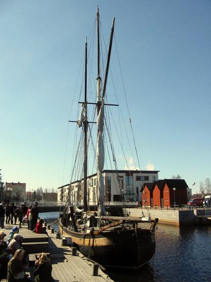 Photo from a spring day in Oulu waterfront, with some people celebrating May Day, sitting by the pier. Docked on the pier is a small old sailboat. A bridge leading to an island with old storehouses and modern apartment buildings can be seen on the background.