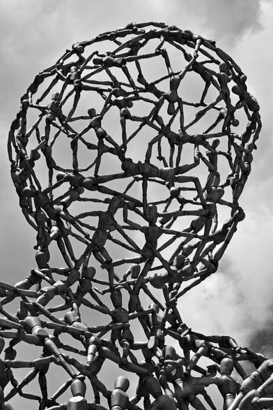 Black and white photo of a section of a large outdoor sculpture composed of hundreds of small metal artist’s mannequins which have been joined together to form the shape of a human head