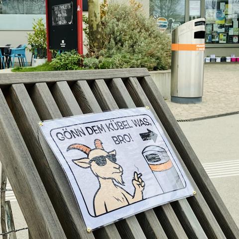 Das Bild zeigt einen der Holzsessel an der Seestadt Lounge. Auf der Lehne des Sessels ist ein laminiertes Blatt mit der Aufschrift "Gönn dem Kübel was, Bro!". Darunter ist eine Illustration einer Ziege mit Sonnenbrille und Zigarette neben einem Mistkübel. Zwischen Text und Illustration ist ein Pfeil. Der Pfeil deutet auf einen Mistkübel der MA48 hinter der Seestadt Lounge zwischen Stadtteilmanagement und See22.