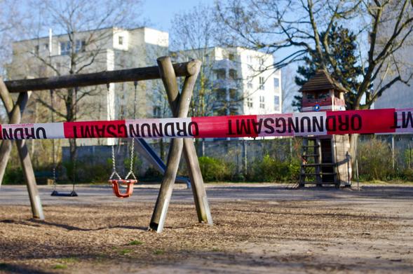 Blick auf einen Spielplatz mit 2 Schaukeln an einem Holzstammgerüst, eine davon einen Kleinkinderschaukel. Weiter hinten eine Rutsche und ein Turm zum Klettern. Im Hintergrund Mietshäuser. Im Vordergrund quer durch das Bild ein rot-weißes Absperrband mit de Aufschrift „Ordnungsamt“. Die Schrift steht auf dem Kopf.