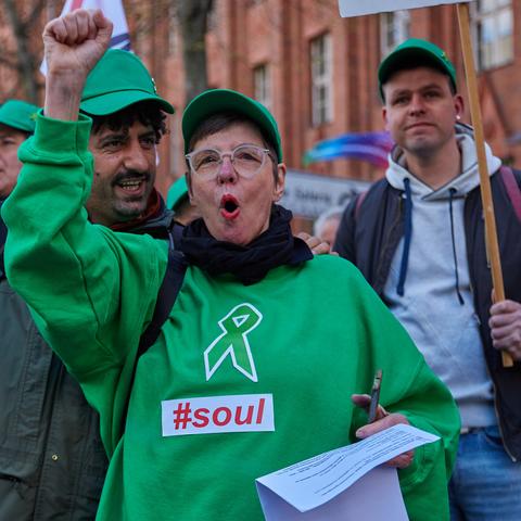 Person in grünem Pullover mit aufgemalter Schleife die die Faust hebt und etwas ruft. Sie und die Menschen hinter ihr tragen auch eine grüne Kappe.