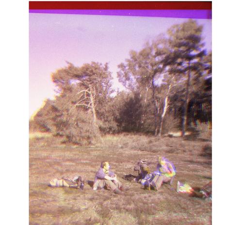 Theo hikers sitting in a sunny field with some pine trees in the background, a few bags scattered around them. It's a colour image from three overlapping layers, red, blue and yellow. Where the layers don't match up, magenta and yellow edges and overalls are visible, a bit like an old and very cheap, badly printed comic
