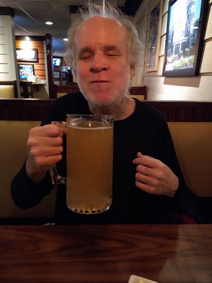 The image depicts a man sitting at a table in a restaurant. He is holding a large glass mug filled with a light amber-colored beverage, possibly beer, given the setting and the mug's size. The man has white hair and a beard, and he is wearing a dark-colored shirt. The background shows part of the restaurant, including wooden booths, a television screen, and a picture frame on the wall. The atmosphere appears casual and relaxed.