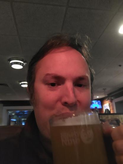 The picture depicts a man who is drinking a beverage. He is holding a mug filled with a light brown liquid, possibly beer. He has a serious expression, with his lips pursed. The man is wearing a black shirt. The setting appears to be a bar or a restaurant. There are two round lamps on the ceiling and a television screen in the background.