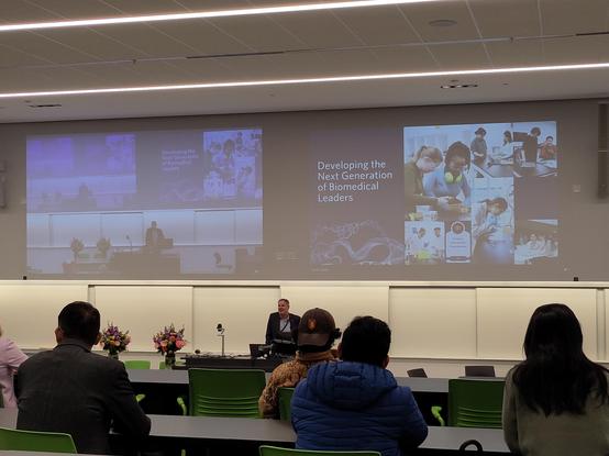 A photograph from the back of a lecture theatre of a speaker giving the welcome to a biomedical engineering research symposium. There is a slide visible with the title "Developing the Next Generation of Biomedical Leaders"