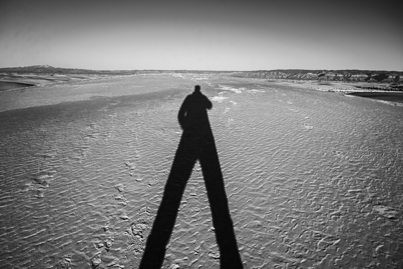 A black and white photo of a long human shadow cast across a rippled sandy desert landscape under a clear sky, with distant mountains along the horizon.