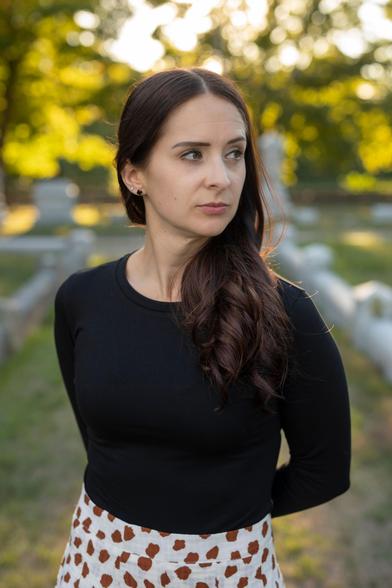 Waist-up color photo of author Christa Carmen, a thin, fair-skinned woman with dark hair and eyes. She wears her hair long and parted in the middle; it falls over one shoulder. She wears a black crewneck knit top, a patterned skirt, and a few pairs of small earrings. She stands among sunlit trees, her body facing the camera as she looks over one shoulder with a sharply focused but inscrutable expression.