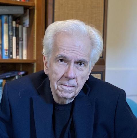 Head-and-shoulders color photo of author Gregory Frost, a fair-skinned man with large brown eyes, white hair parted on the side, and a long, thin face. He wears a black blazer over a black crewneck shirt and sits in front of a wooden bookcase full of books. He looks straight into the camera with an intense expression.