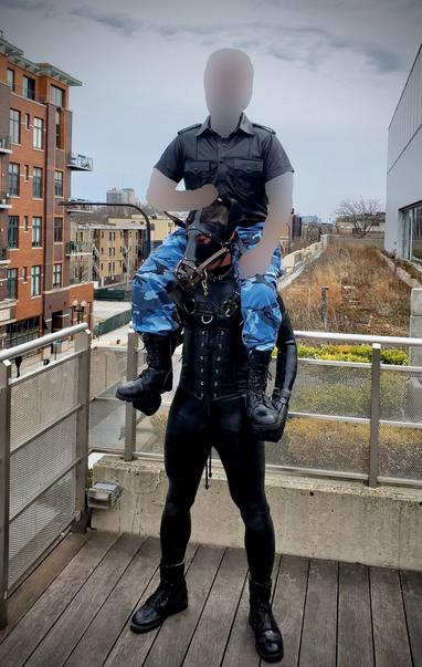 A person in a full black silicone bodysuit is styled as a human horse, complete with a sleek leather corset featuring prominent lace-up details. The figure stands tall in glossy black boots on wooden decking, embodying the aesthetic of human pony play.

Mounted atop this impressive pony sits a rider wearing a military-inspired black shirt with epaulets and shoulder details, paired with distinctive blue camouflage cargo pants. The contrast between the structured military-style top and the tactical camo bottoms creates a commanding presence. The rider's combat boots rest in the pony’s hoofs, which are wrapped in leather puppy mitts that reflect the pony’s hybrid identity as a ponyhound.

The scene unfolds against a dramatic urban backdrop of brick apartment buildings and city architecture. The overcast sky casts a moody atmosphere over the rooftop setting, where metal railings frame the edge of the balcony. In the background, patches of early spring vegetation can be seen on an adjacent rooftop garden area, adding a touch of nature to the metropolitan environment.

This powerful image captures the intersection of alternative lifestyle expression and urban living, showcasing the artistry and dedication involved in human pony play relationships. The composition balances intimate personal expression with the expansive cityscape beyond, creating a compelling visual narrative about community, identity, and connection.

#HumanPonyPlay #LeatherCommunity #AlternativeLifestyle #UrbanSce