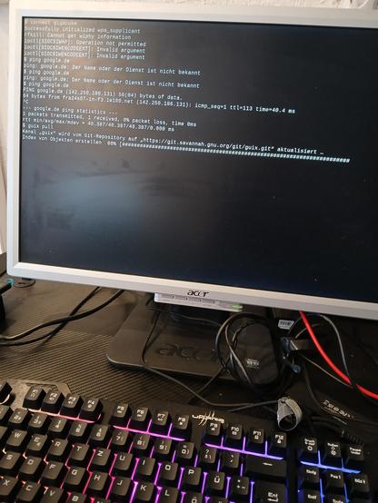 Ein Monitor, davor eine RGB beleuchtete Tastatur und diverse Kabel. Auf dem Monitor ist eine shell zu sehen die ein "guix pull" ausführt
