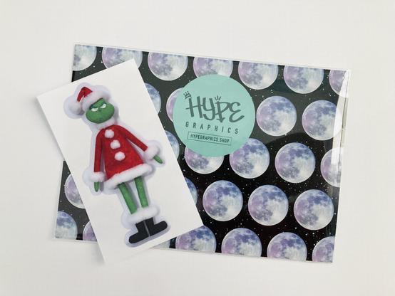 A Grinch fabric doll kiss cut sticker laying on marketing materials from Hype Graphics (an Etsy store)
