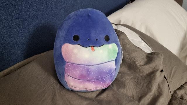 'Squishmallow' soft toy in the shape of a fat snake with a starry belly, sitting on a grey pillow.