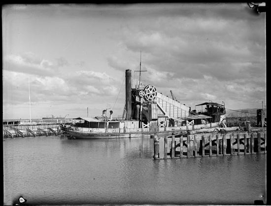 Black-and-white photo: Black and white image of the dredge KORUA. ca 1940s. Photographer, Stanley Vincent. Description: A view of the starboard side of a rather dilapidated, single-funnelled dredge tied up at wooden docks. Citation: Tairawhiti Museum, Accession No. 2012.80.8. https://collection.tairawhitimuseum.org.nz/objects/33703/glass-plate-negative