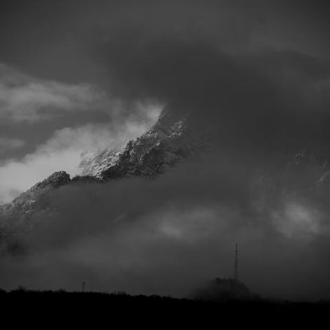 Photo noir et blanc de montagne.
Une montagne très abrupte, partiellement masquée par des lambeaux de nuage, recouverte par un saupoudrage neigeux.
Au premier plan, en ombre chinoise, une colline surmontée d'un pylône et d'une tour télécom.