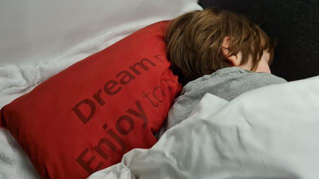 Detail of the cabin of the ÖBB Nightjet night train. A young cute child sleeps, his head resting on a red pillow branded with the ÖBB Nightjet logo and the tagline “Dream now. Enjoy tomorrow”.