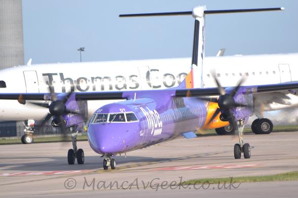 Almost head-on shot of a very purple, high-winged, twin propellor-engined airliner, taxiing from right to left, pointing to just to the left of the camera.
The body is purple, with large, billboard-style "FlyBe" titles on the forward fuselage, just aft of the forward doors.
There is a yellow and red band round the rear fuselage, and a white tail with barely distinguishable "FlyBe" titles.
The engine pods on the wings are the same purple, with white text "Faster than road or rail".
Grey concrete taxiway fills the foreground, while a white, twin engined jet airliner  with yellow engine pods under the wings and grey "Thomas Cook" titles on the side of the white forward fuselage, taxis from right to left in the background, under grey-blue skies.