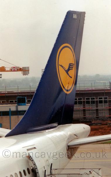 Close-up shot of the blue tail of a white jet airliner, parked facing to the left.
The tail has a large yellow circle, with a stylised blue flying bird in the middle.
A brown building stretches across the background, with the balance boom of a large yellow crane in the distance on the left of the frame.
Grey sky fills the rest of the frame.