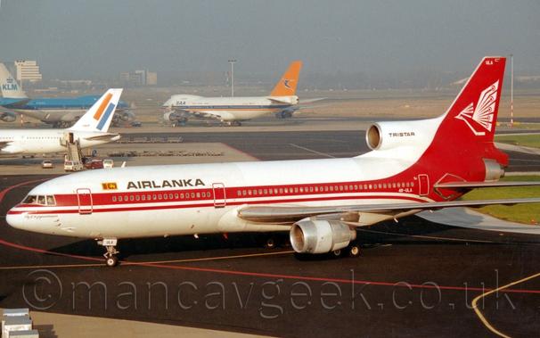 Side view of a white, 3 engined jet airliner with red piping on the body and a red tail, taxiing from right to left at a busy airport.
There is a thick red stripe running along the body, sweeping up and down to fill the rear fuselage and tail
The tail has a stylised white image of a bird with spread wings.
There are black "Air Lanka" titles on the upper forward fuselage.
Black concrete apron fills the foreground, with red and yellow markings for planes to follow.
3 jet airliners are visible in the background - the rear fuselage and tail of an all-white jet airliner with orange and blue diagonal stripes on the tail on the left of the frame, a very large, 4 engined blue and white jet airliner with blue "KLM" titles on the white tail just behind that, and a white, 4 engined jet airliner with a blue and orange stripe running along the body and an orange and blue tail in the distance in the middle.
Tall buildings mark the periphery of the airport, slowly vanishing in to grey haze.