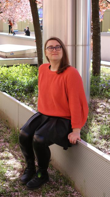Me with long hair, wearing an orange knitted sweater and fake leather skirt, leggings and boots. I am sitting on the edge of a flowerbed and there is a big metal cylinder behind me