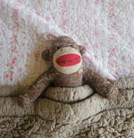 Sock Monkey dog toy tucked into a tan fuzzy blanket.  He is laying on a pastel pink/white/green flowered quilt.
