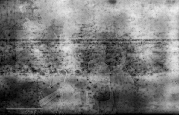 Very old black and white photo. Foggy and spotted, with markings from the backing paper showing, you can barely make out some people in old fashioned clothes and hairdos.