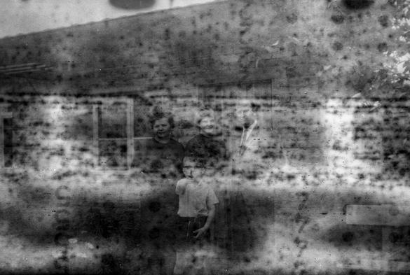 Very old black and white photo. Foggy and spotted, with markings from the backing paper showing, you can barely make out some people in old fashioned clothes and hairdos.