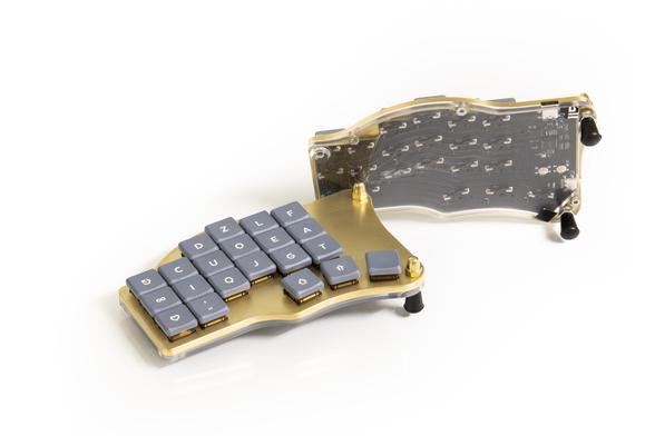 A splitkeyboard made of brass. The two halfes are shown, one from the upper side, showing the keys, one from the back showing the pint through the policarbonate back cover.