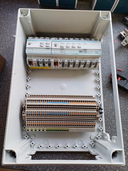 A electrical enclosure with DIN rail-mounted KNX actuators at the top. Below them are terminal blocks for wiring connections. The enclosure is placed on a workbench.