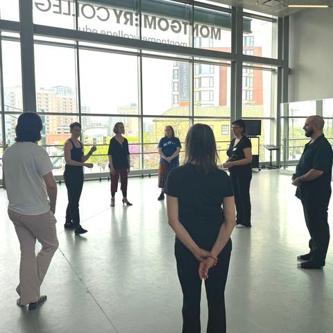 7 people stand in a circle in front of a large window. One holds up their hands, explaining.