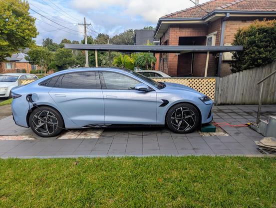 Side on photo of a BYD Seal electric car parked in a driveway. There is a cable running from the charging port at the back of the car to a green box on the ground by the front of the car, and two cables running from that box out of shot.